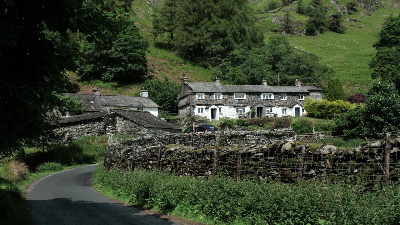 Static shot of traditional stone cottages beside a winding country lane in the Lake District, surrounded by dry stone walls, green hills, and peaceful rural scenery