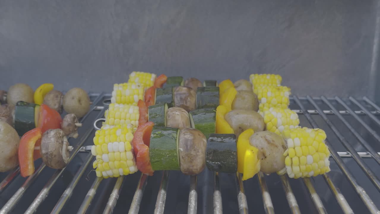 Closeup shot of vegetable skewers with corn, pepper, zucchini, potatoes and mushrooms are being cooked on a stainless steel bbq and brushed with oil. Smoke rises from the grill