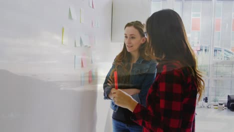 Composite-video-of-a-beach-over-two-diverse-female-colleagues-discussing-over-memo-notes-at-office