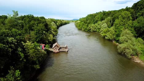 Paisaje-Aéreo-Tiro-Descendente-De-Un-Molino-De-Barcos-Sobre-Los-Bosques-Del-Río-Mura-En-Ambos-Lados-Cielo-Nublado-Austria-Eslovenia-Europa