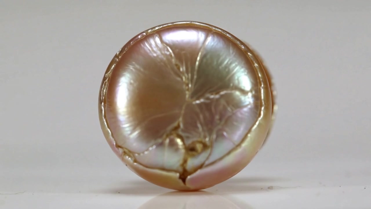 Perla iridescente che ruota da un'angolazione, rivelando segmenti di petali e crepe radiali su un tavolo