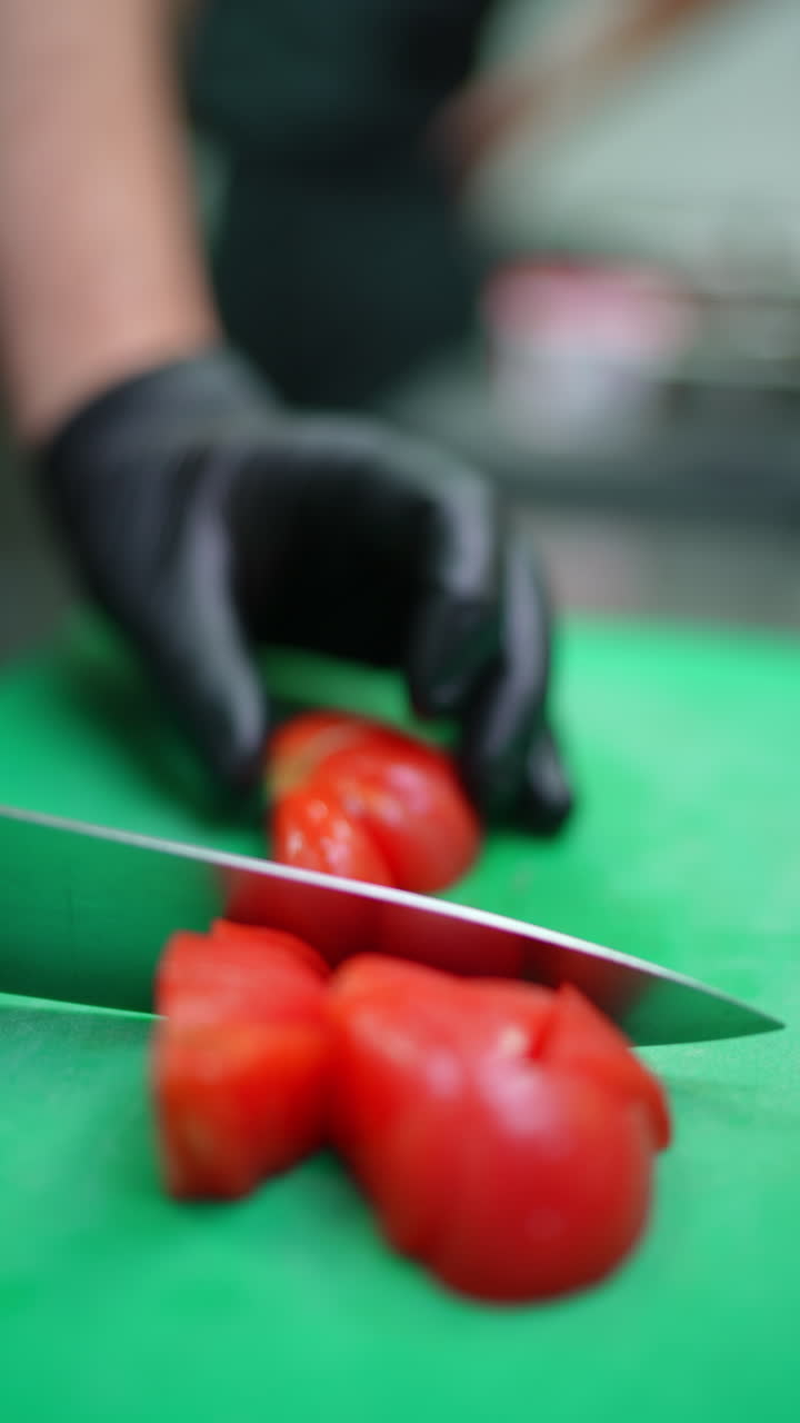 Vertical footage Professional chef wearing black protective gloves carefully slicing ripe red tomatoes on green cutting board using sharp knife during food preparation in commercial kitchen
