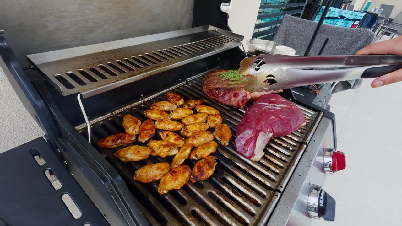 A Person Holding Tongs Grilling Chicken Wings And Steak In Summer. High Angle Shot