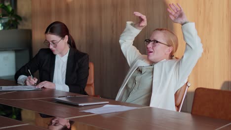 una chica rubia segura y feliz con gafas y un traje de negocios blanco cierra su portátil y se apoya en el respaldo de su silla durante el éxito y la victoria al final de la jornada laboral en una oficina moderna