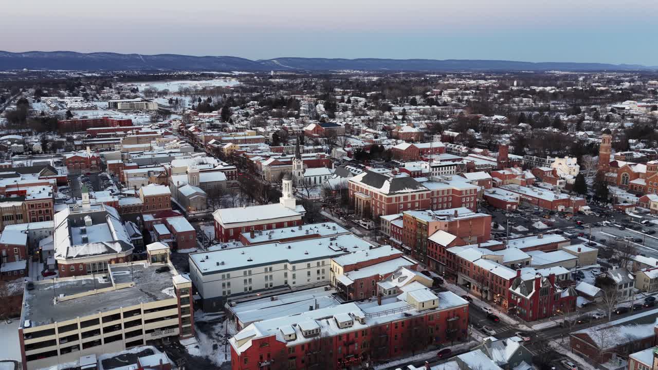 Luftdroneudsigt over en lille amerikansk by ved solnedgang om vinteren, med snedækkede hustage, gader med let trafik og historiske bygninger, der strækker sig mod fjerne bakker. Fredfyldt juletid