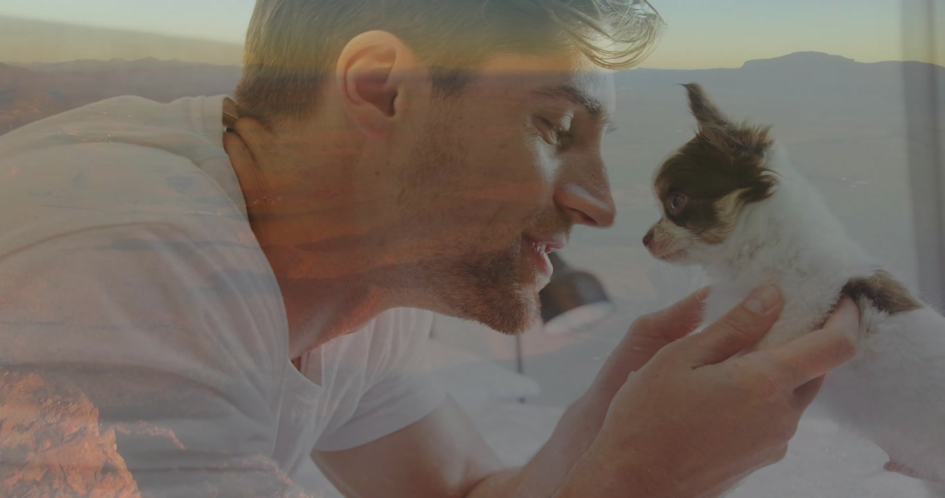 Mid-adult man gripping puppy legs then kissing nose and smiling while promoting pet care in bedroom