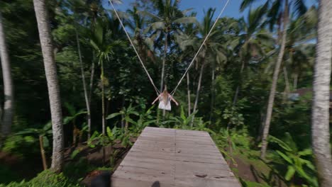 blond woman in a summer dress swinging on a long swing set in the tropical nature of bali