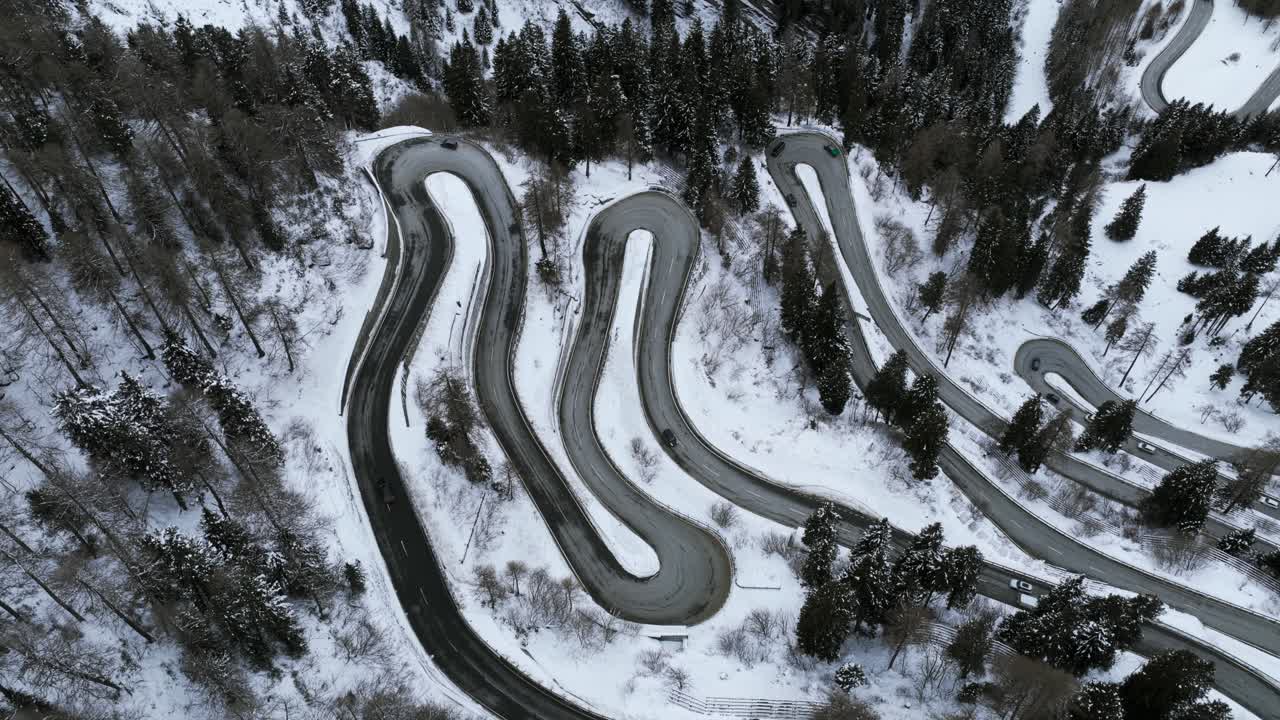 Serpentine mountain road with hairpin turns through a snow-covered forest in the Swiss Alps. Aerial drone ascending, top-down view