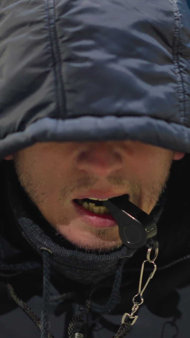 primer plano de un hombre con una capucha que cubre su cara, sosteniendo un silbato en la boca, el fondo está débilmente iluminado, con una vista borrosa de un edificio y un campo