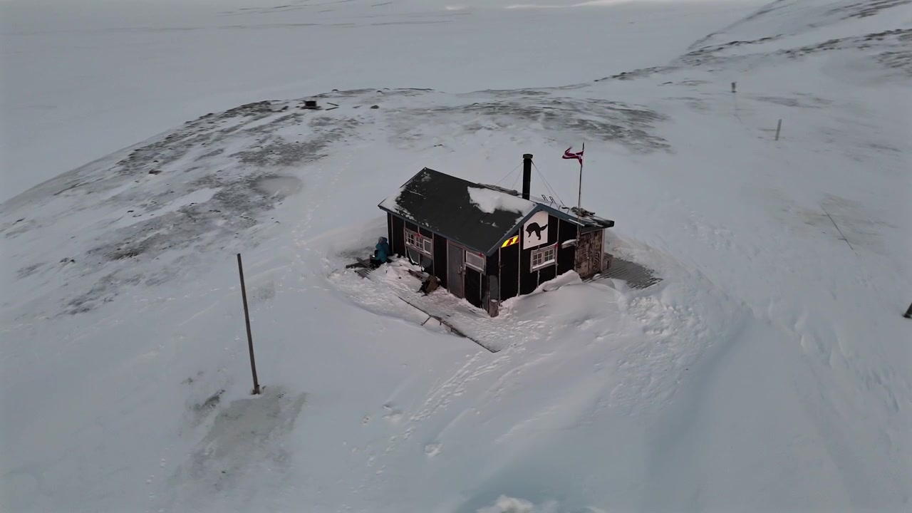 Aerial drone orbit shot around a small snow covered cabin with a smoking chimney, warning sign, and a Danish flag flying in Svalbard