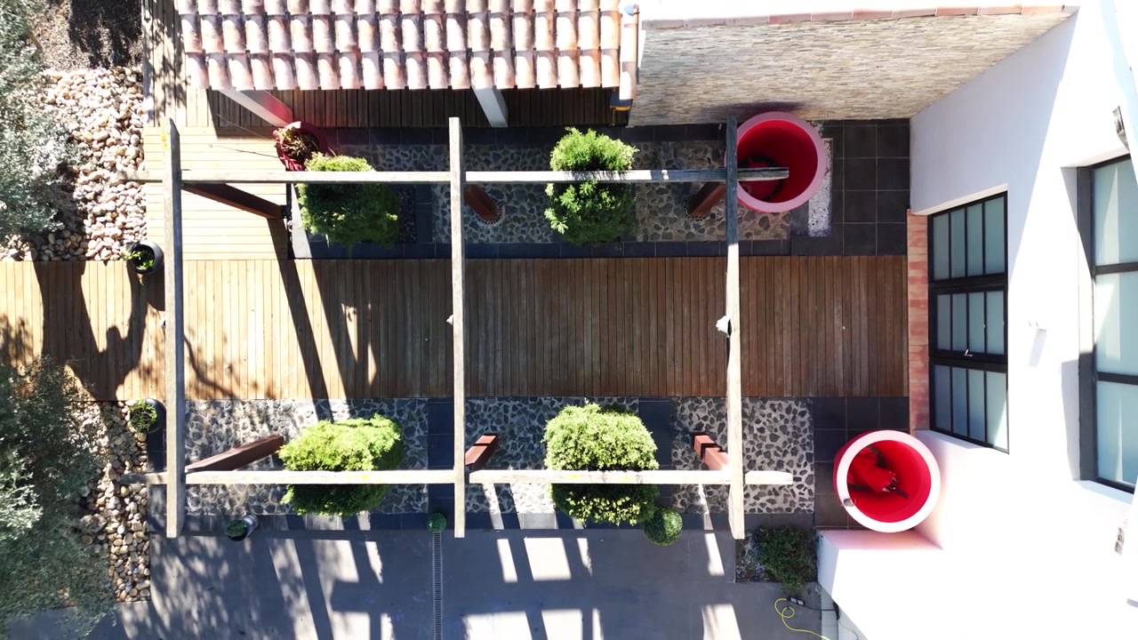 Ripresa verticale con drone che si eleva sopra una passerella in legno, arbusti verdi e un'area patio moderna.