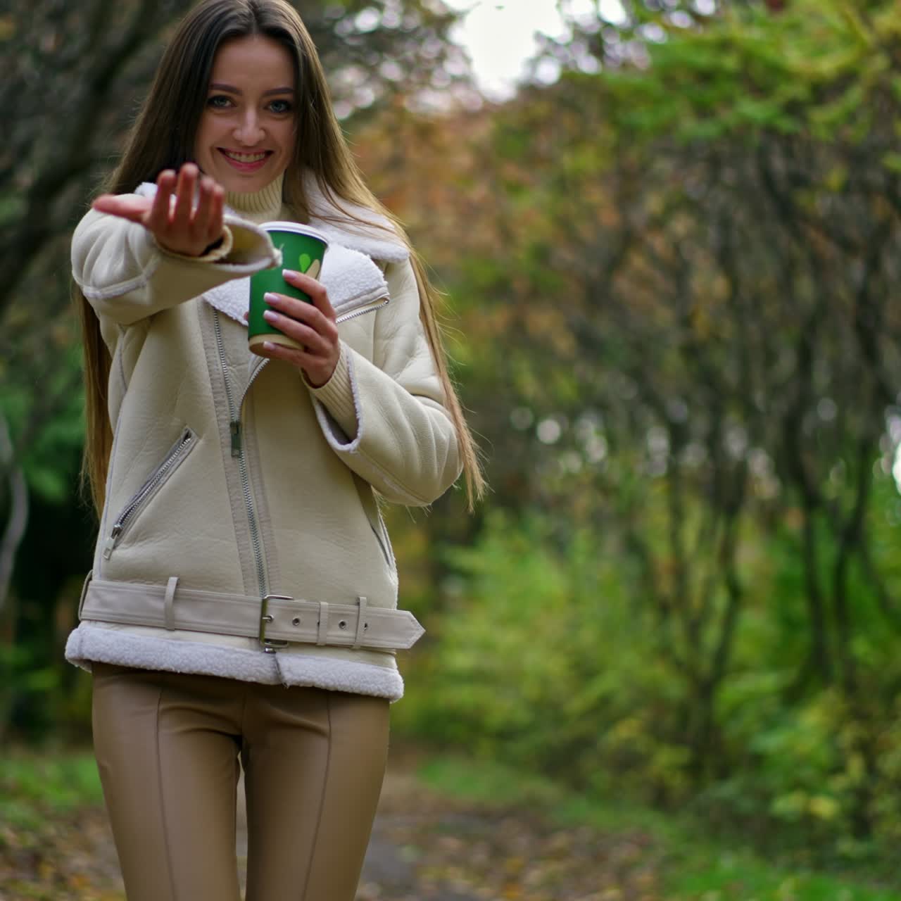 Una signora bruna, sorridente e con i capelli lunghi, passeggia nel parco con un bicchiere di carta in mano. La signora sorride alla telecamera e gesticola chiamando con la mano. Parco autunnale sullo sfondo
