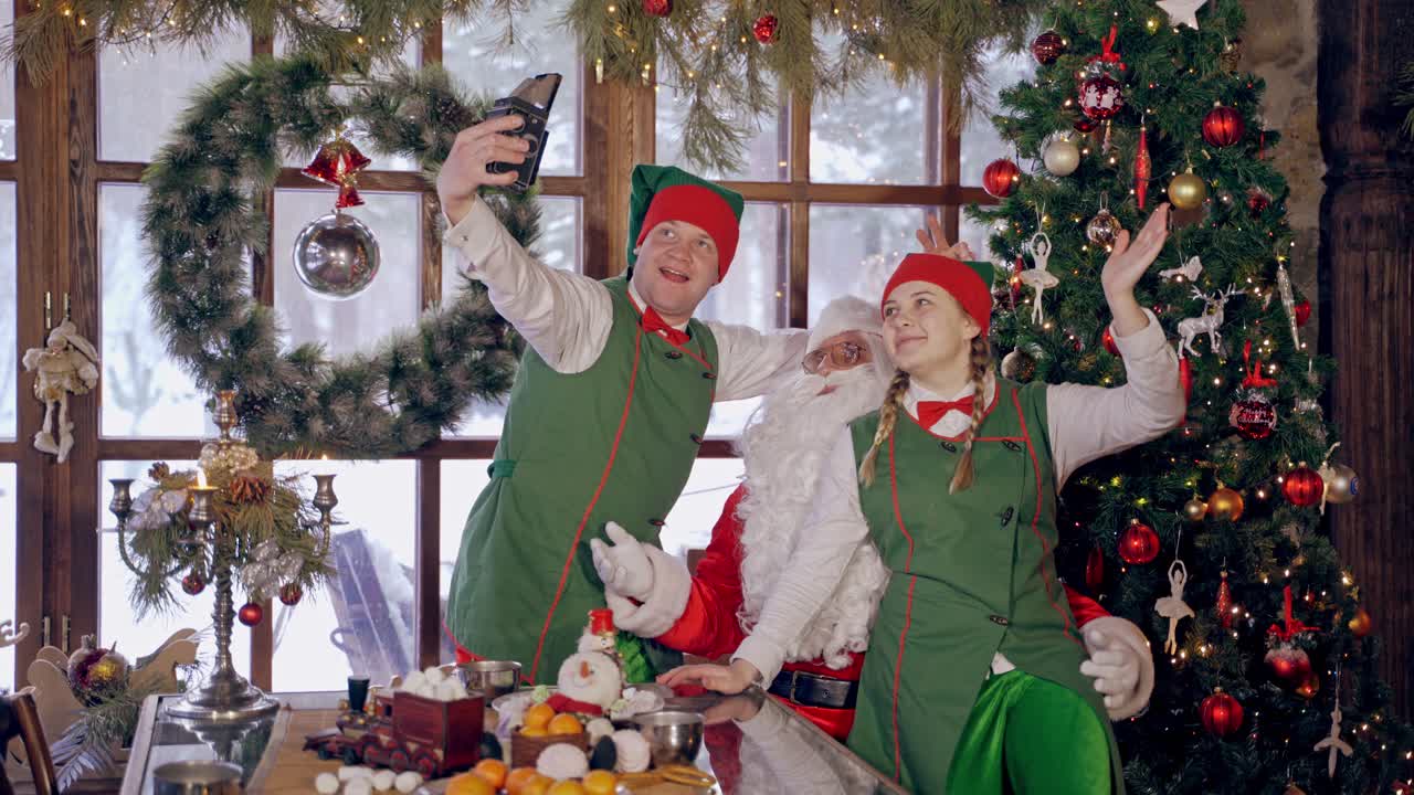 Fairy elves photographing with Santa Claus. Santa with white beard and elves in green costumes doing selfies on Christmas holidays. Christmas decorations.