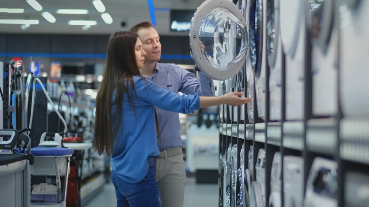 A handsome man and a woman choose a washing machine to buy open the door and inspect the coating of the drum and the quality of the product. Woman looking inside washing machine