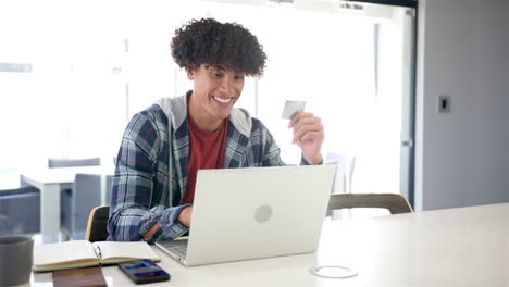 un joven biracial está haciendo compras en línea, usando su portátil y tarjeta de crédito con espacio para copiar