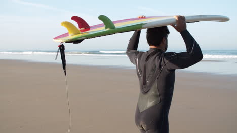 vista posterior de un surfista masculino con pierna protésica caminando en la playa con tabla de surf en la cabeza