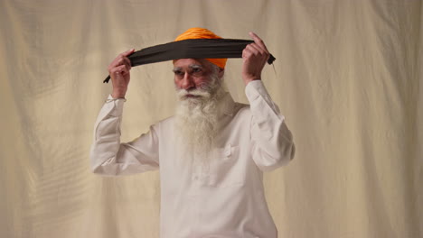 studio shot of senior sikh man with beard tying fabric for turban against plain background as sequence part 1 of 3