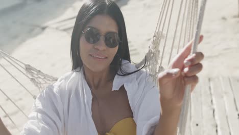 retrato de una feliz mujer hispana con gafas de sol tendida en una hamaca en una playa soleada, en cámara lenta.