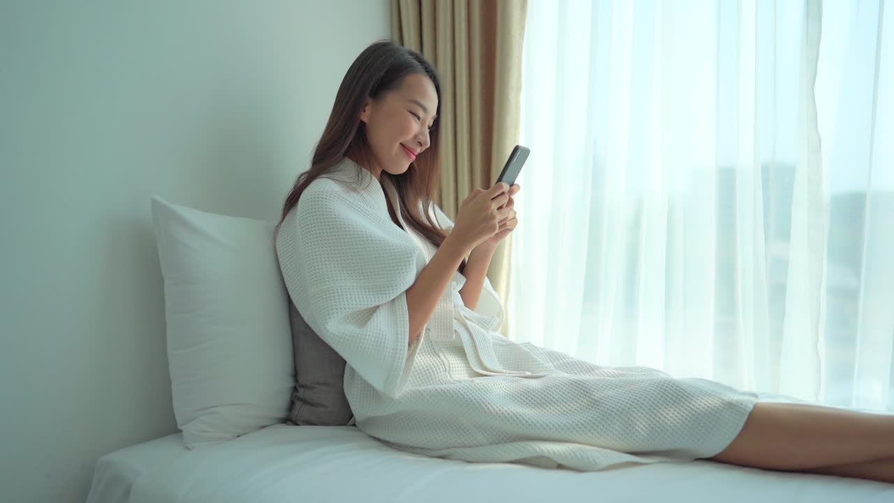 A young woman in a resort bathrobe is sitting up in bad while texting on her smartphone,