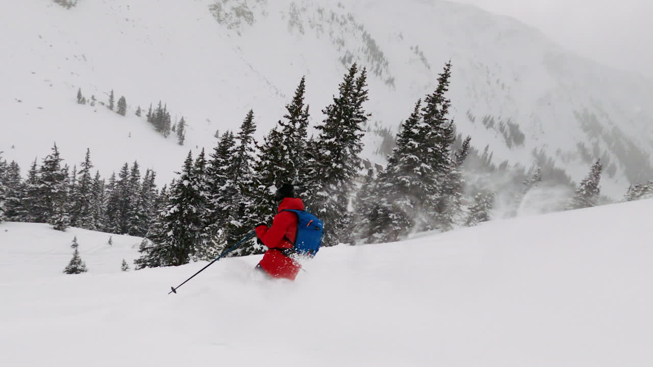 Scialpinista uomo con giacca rossa Northface che pratica sci fuori pista su neve fresca e polverosa giù per il versante della montagna, ripresa GoPro. Sfondo con paesaggio montano del Colorado, Coon Hill, Tunnel Eisenhower, I-70 e Continental Divide.