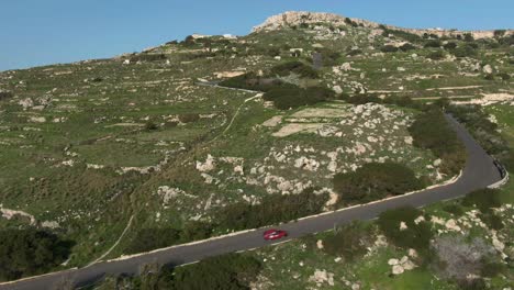 red car driving on the road uphill with lush rocky meadows on the sides - aerial shot