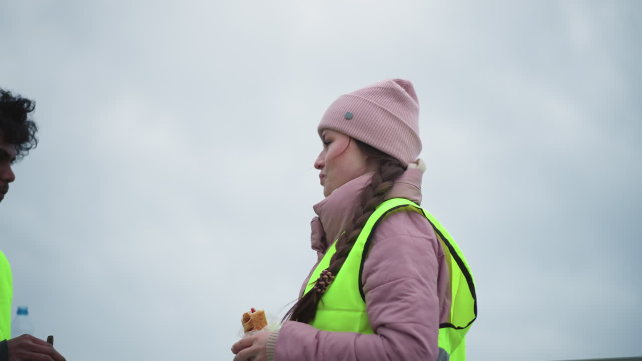 Woman in pink beanie and puffy jacket with reflective safety vest holding wrap sandwich, speaking outdoors on cloudy cold day, long braid over shoulder, water bottle in background