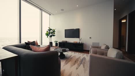 wide push in of a living room setup showcasing a small tree, glass coffee table, couches, and tv, next to large windows letting in natural sunlight