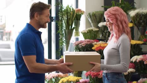 a man hands over a brown box of fresh flowers to a smiling woman in a lively floral shop. the shop showcases colorful arrangements, creating a cheerful atmosphere.