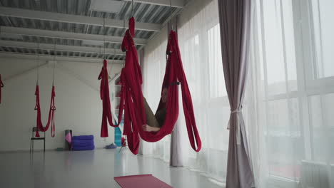 bright yoga studio with floor to ceiling windows and sheer curtains as mindfulness practitioner lies comfortably in red silk hammock swinging gently, conveying calm balance focus wellness