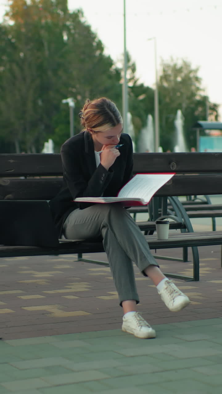 Studentessa concentrata seduta all'aperto su una panchina di legno, che legge un libro con un laptop accanto e una tazza di caffè in un ambiente urbano circondata da panchine vuote, terreno pavimentato, alberi e una tenue luce serale