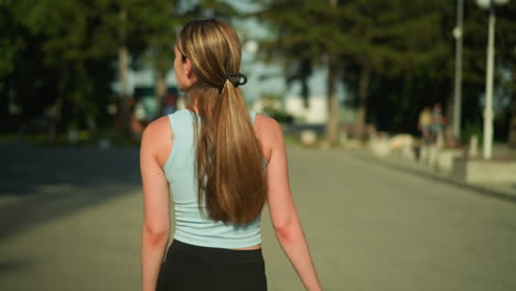 back view of young lady in athletic wear walking on interlocked path with her hair tied back, glancing side to side, sunlight enhances her movement while people are blurred in the background