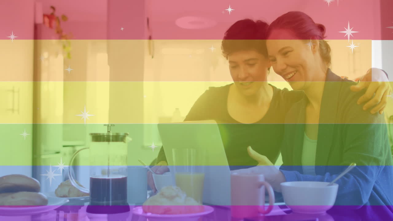 Animation of pride text, rainbow stripes over happy caucasian lesbian couple using tablet at table