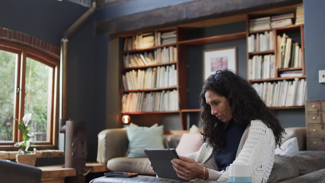 mujer biracial sentada en el sofá y usando la tableta en casa, cámara lenta con espacio de copia
