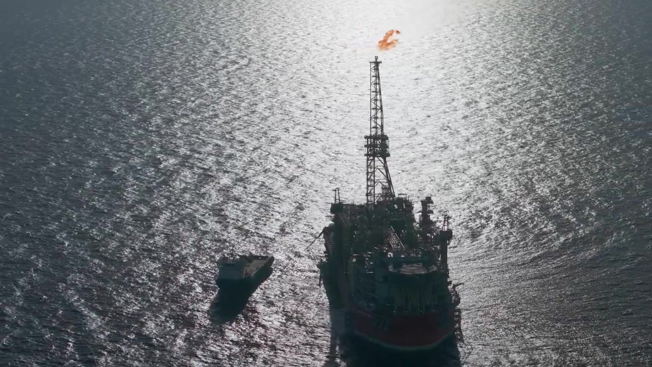 Aerial drone shot of a support boat navigating near a large floating production storage vessel with a burning flare stack against shimmering sea water.