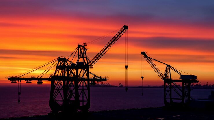 cargo cranes standing tall and silhouetted against a breathtaking sunset sky in an industrial port area, with metallic structures of the cranes and a cloudy sky displaying warm and cool-ton.