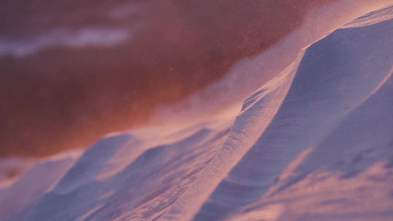 Slowmotion close up of snow blowing in the wind as sunset lights the scenery orange in the background