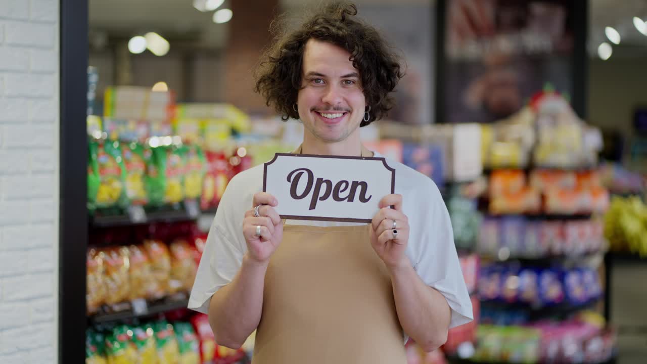 Portrait of a happy brunette guy with curly hair who holds in his hands a sign with the inscription open after the opening of the supermarket