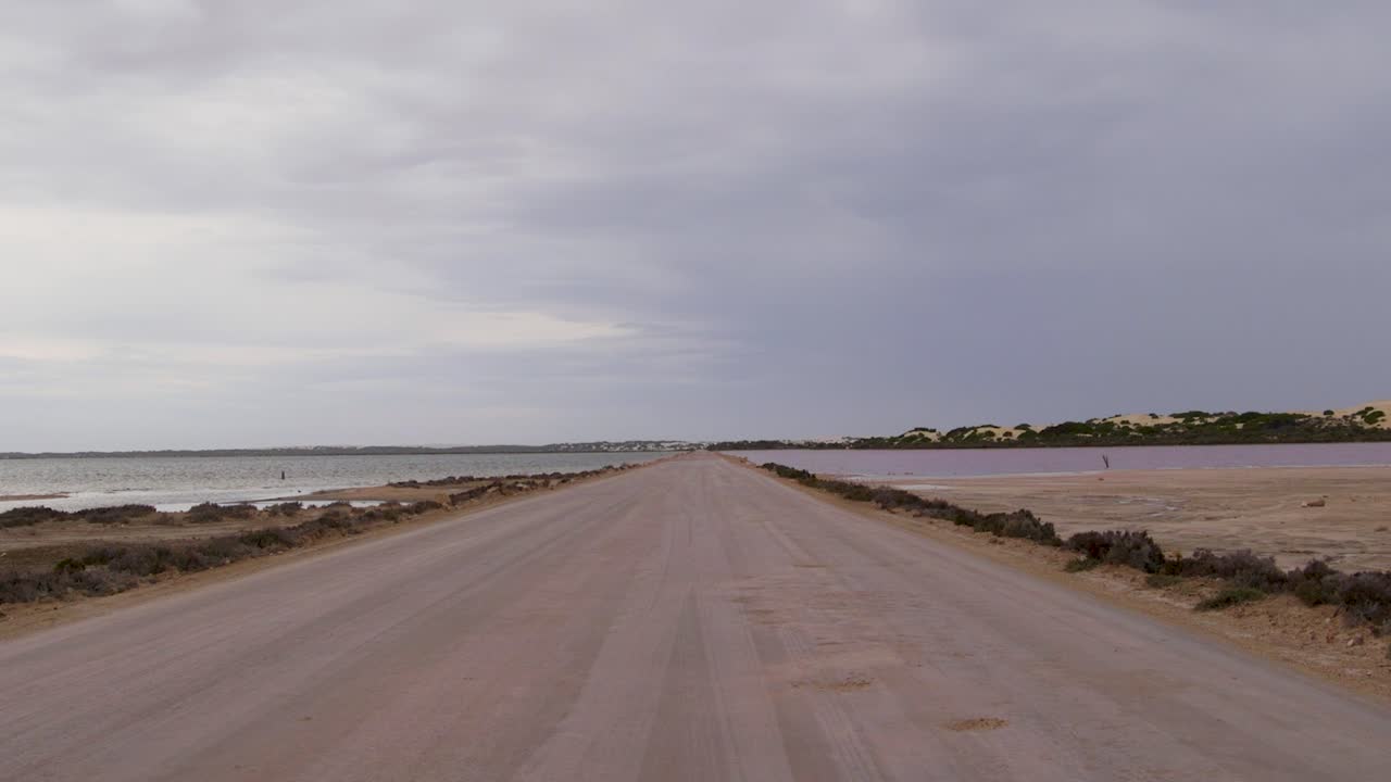 Una carretera larga y recta se extiende entre un lago salado rosa y el océano bajo un cielo nublado.