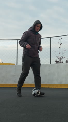 hombre practicando ejercicios de fútbol en una arena deportiva, demostrando control de la pelota y enfoque bajo la luz del día suave, el fondo incluye edificios residenciales urbanos, postes de luz, cercas de malla y un poste de portería