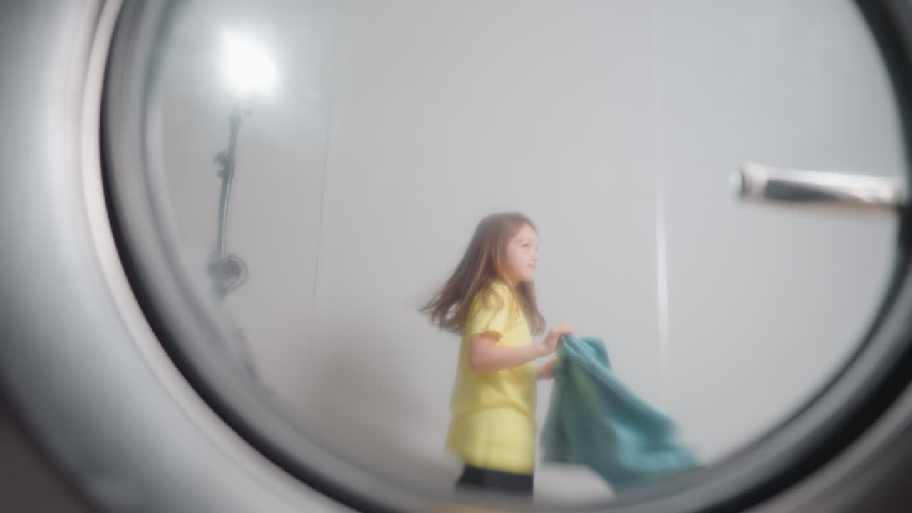 Industrial washer door view shows little girl holding blue towel, dancing excited, playful movement seen through transparent window, laundry setting, joy, clean fabrics, modern laundromat scene