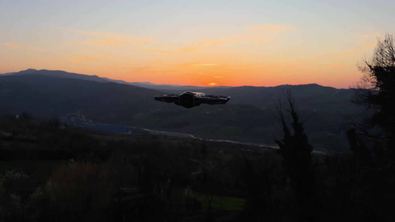 Drone FPV in volo stazionario nell'aria dorata contro un vibrante cielo arancione al tramonto sopra una cresta montuosa.