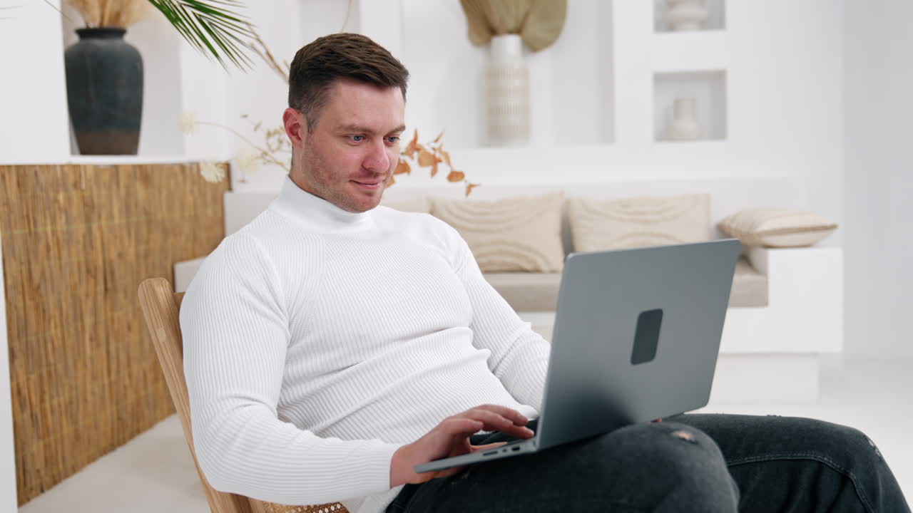 Dark-haired fit man sits working at laptop. Man looks at camera smiling. Work in cozy home atmosphere.