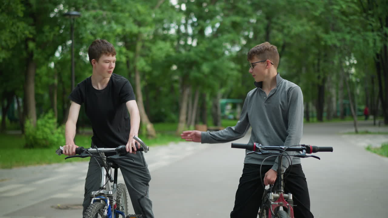 두 명의 친구가 자전거를 타고, 회색 을 입은 사람은 친구가 그를 환영하기 위해 올라오는 것을 기다리고, 회색을 입은 소년은 손목 부히기 위해 손을 고, 배경에는 흐릿한 푸른 나무가 있습니다.