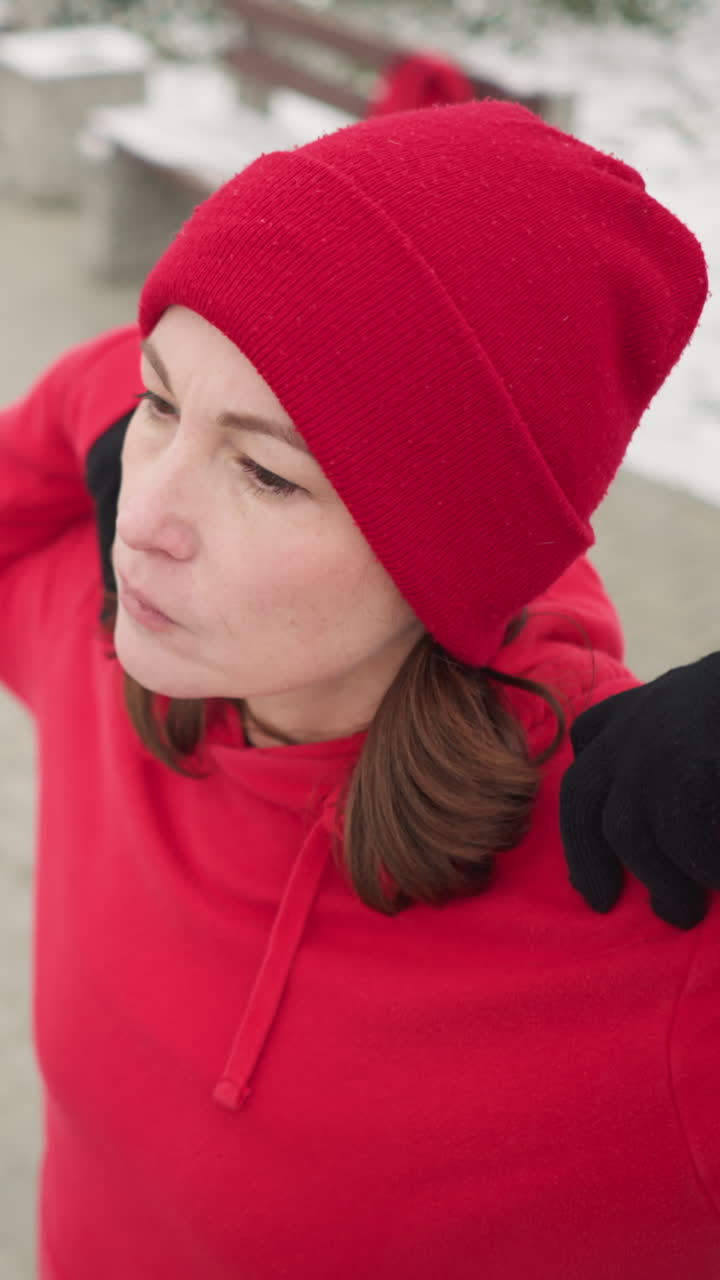 dama realizando estiramiento de manos girando el brazo al aire libre en la rutina de fitness de invierno, vestida con ropa atlética, suelo cubierto de nieve, árboles helados, bancos en un entorno de parque sereno