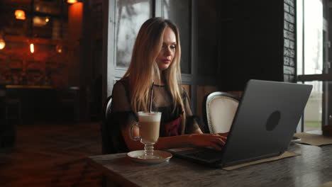 Side-view.-Young-business-woman-sitting-at-table-and-taking-notes-in-notebook.On-table-is-laptop,-smartphone-and-cup-of-coffee.On-computer-screen-graphics-and-charts.-Student-learning-online.-Blogger.