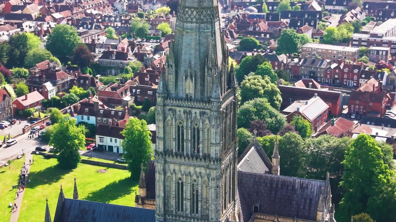 Drohnen-Luftaufnahme der Kathedrale von Salisbury in Wiltshire, England, die den mittelalterlichen gotischen Kirchturm zeigt, der an einem hellen Sonnentag über historischen Dächern, Bäumen, Straßen und Stadtgebäuden aufragt.