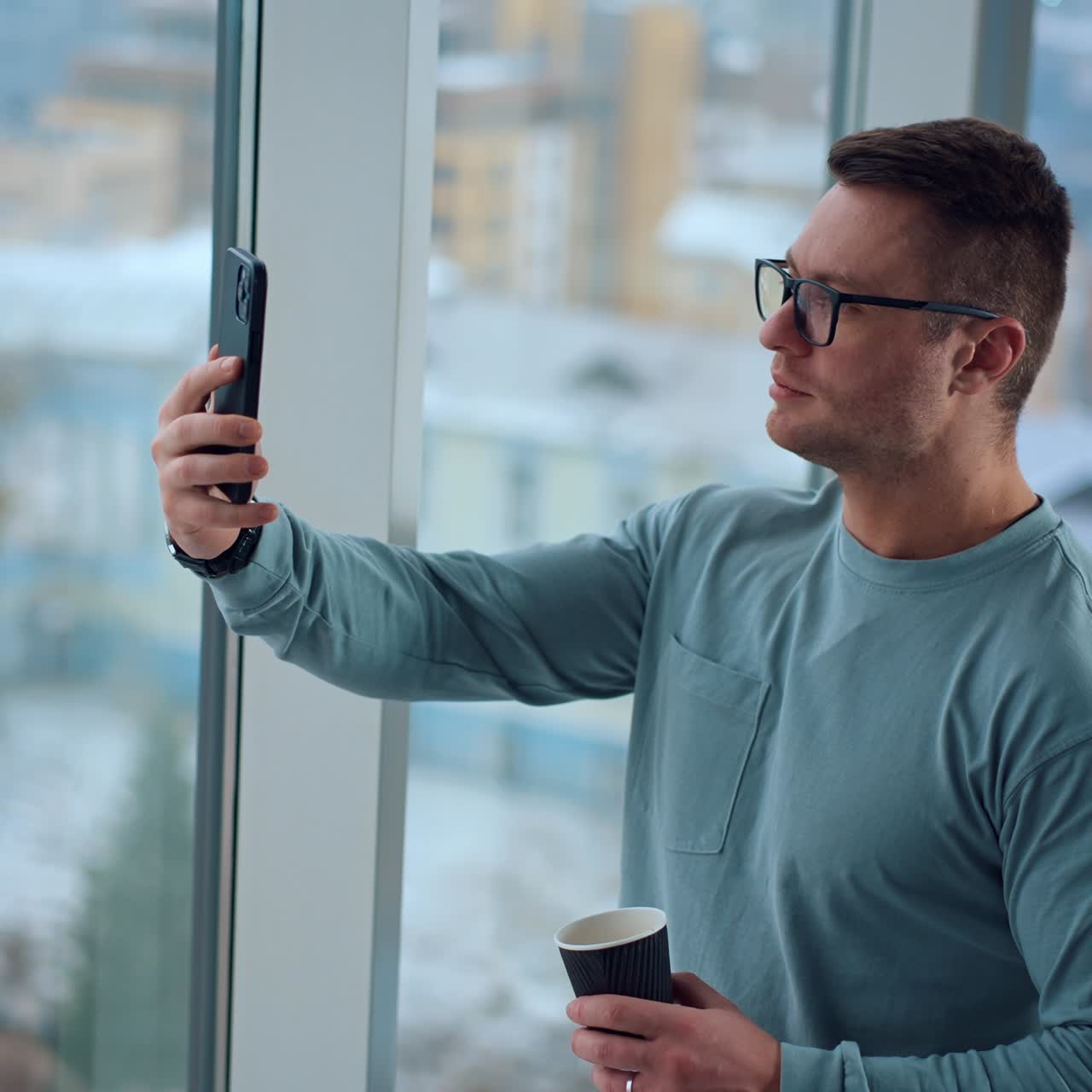 Man holding a phone in front of him taking video or selfie. Strong Caucasian male with a paper cup in hand standing indoors at the window