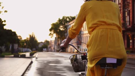 back view of and unrecognizable woman in long yellow dress riding a city bicycle with a basket and flowers in the city center