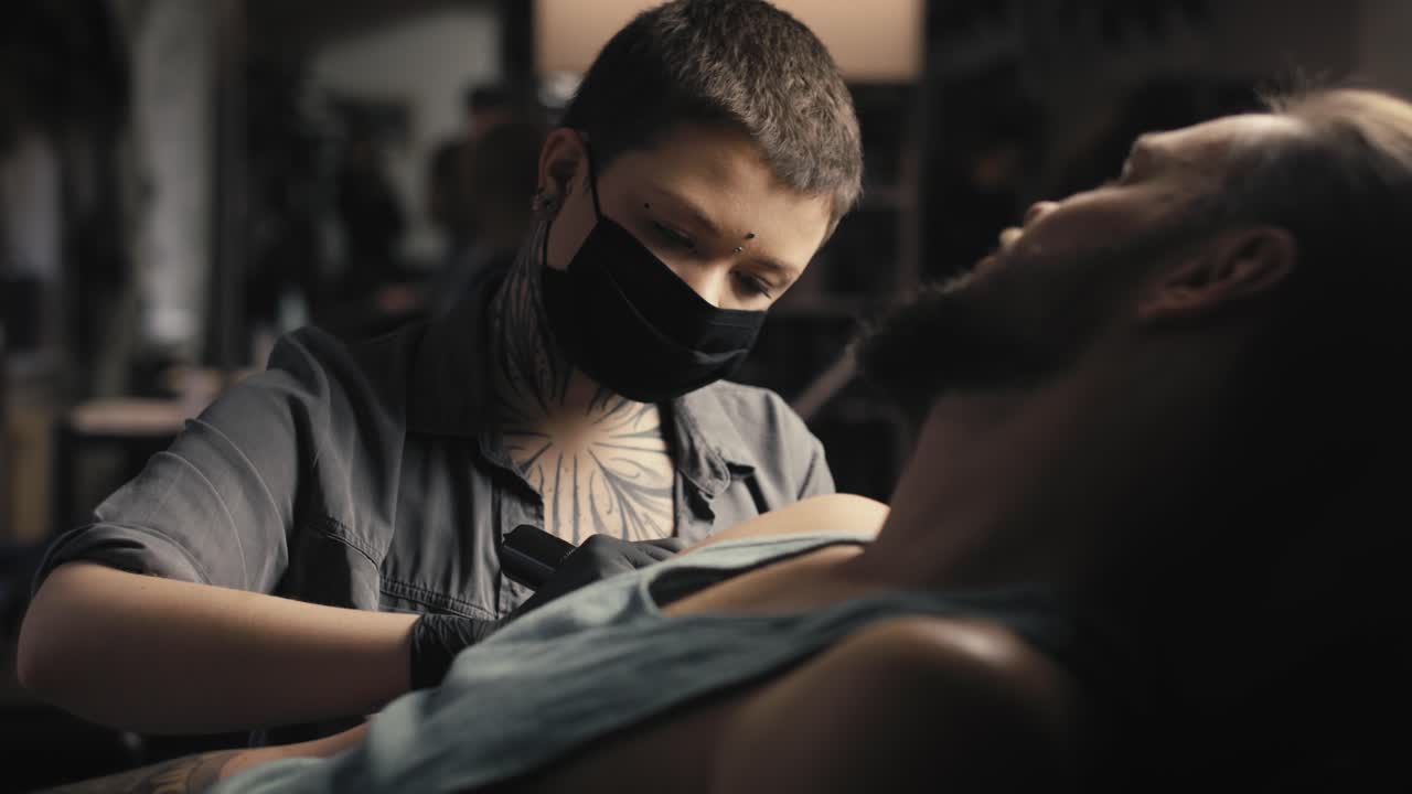 얼굴 마스크를 쓴 백인 여성이 고객의 팔에 문신을 하고 있습니다.