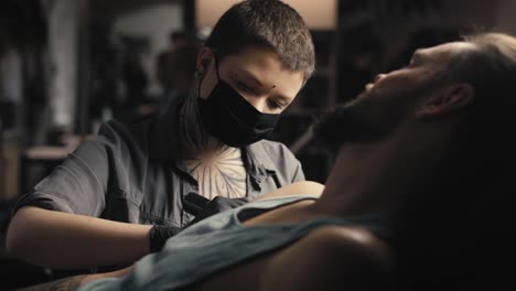 Caucasian-woman-in-face-mask-tattooing-arms-of-her-customer.